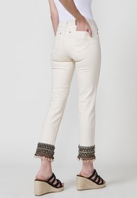 Jeans de denim color crema con adornos de cuentas decorativos en el tobillo. Presenta dos bolsillos traseros y una silueta ajustada. Combinados con sandalias de cuña negras.