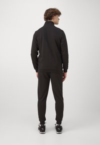 Tuta nera con giacca zip-up e polsini elasticizzati, realizzata in misto cotone. Texture sottile, colletto a costine e design aderente.