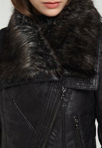 Veste en similicuir noir avec un large col en fourrure, une fermeture éclair inclinée et des détails texturés côtelés sur le corps. Dispose de deux poches latérales zippées.