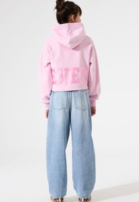 Sudadera rosa corta con capucha y la palabra "EVER" en letras texturizadas en la espalda, combinada con unos pantalones vaqueros anchos de mezclilla azul clara.