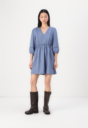 Robe en chambray bleu avec décolleté en V, manches bouffantes et taille cintrée, associée à des bottes en cuir marron à hauteur de genoux avec des accents de boucles.