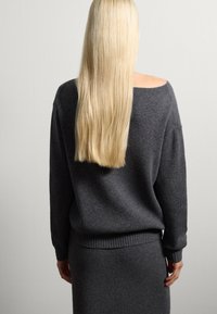 Donna con lunghi capelli biondi indossa un maglione grigio scuro senza spalle e una gonna abbinata, in piedi contro un muro chiaro, di spalle alla macchina fotografica.