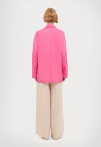 Veste rose à manches longues avec une finition lisse et brillante, portée sur un pantalon beige à jambes larges, vue de dos. Col simple et coupe droite.