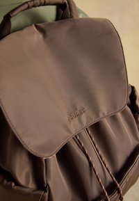 Sac à dos en tissu marron avec fermeture par cordon, rabat supérieur, poignée de transport et logo "JOULES" brodé sur le rabat.
