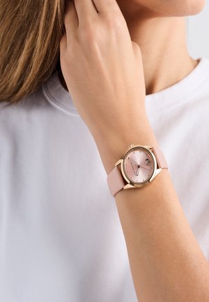 Ümmargune kell roosa kuldse korpuse, roosa diali ja heledast roosa silikoonist rihmaga. Sisaldab tunni-markereid ja väikest kroonit külgedel.