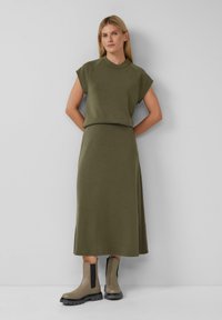 Olijfgroen tweedelig outfit bestaande uit een top met korte mouwen en een hoge halslijn, gecombineerd met een bijpassende lange rok. Gecombineerd met chunky enkellaarsjes.
