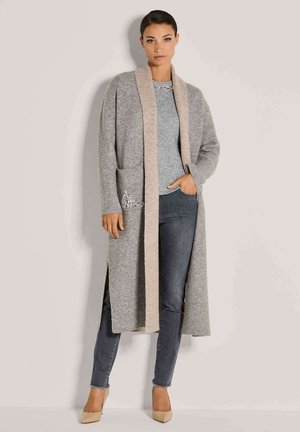 Frau in einer langen grauen Strickjacke mit beigem Rand über einem grauen Pullover, dunkelblauen Skinny-Jeans und beigen High Heels, die vor einem schlichten Hintergrund steht.