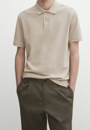 Uomo che indossa una polo beige a maniche corte infilata leggermente in pantaloni verde oliva con le mani in tasca su sfondo semplice.