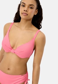 Mujer con bikini rosa texturizado, con parte superior anudada y parte inferior con lazo lateral, de pie frente a un fondo blanco, mirando hacia adelante.