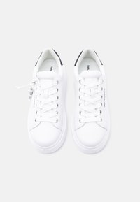 Ζευγάρι λευκά παπούτσια χαμηλής κορυφής με λευκά κορδόνια, μαύρες ταμπέλες στη φτέρνα και ένα μικρό ασημένιο γούρι στο κορδόνι του αριστερού παπουτσιού.