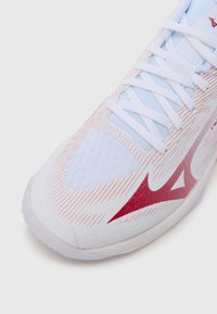 Λευκό αθλητικό παπούτσι με κόκκινες και λευκές ρίγες, ύφασμα από δί�χτυ, στρογγυλή μύτη και επίπεδα κορδόνια. Το λογότυπο είναι ορατό στο πλάι.