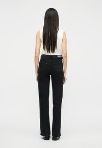 Γυναίκα με μακριά σκούρα μαλλιά που φοράει λευκό αμάνικο τοπ και μαύρο τζιν ψηλόμεσο, στέκεται γυρισμένη πλάτη σε απλό φόντο.