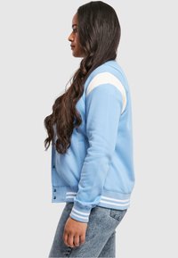 Giacca varsity di colore azzurro chiaro con strisce bianche sulle spalle, polsini a coste e bottoni. Indossata sopra jeans in denim; presenta una vestibilità rilassata.