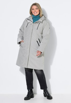 Donna sorridente che indossa un cappotto lungo con cappuccio grigio chiaro, un dolcevita turchese, pantaloni neri con glitter e stivaletti neri, in piedi su sfondo bianco.