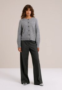 Siv volnen kardigan s krožnim izrezom in šestimi temnimi gumbi, v kombinaciji s prostornimi hlačami z vzorcem črt in srebrnimi čevlji.