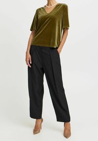 Top verde oliva in velluto a V abbinato a pantaloni neri plissettati e scarpe con tacco nudo; texture morbida e vestibilità rilassata.