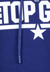 Sininen huppari, jossa on valkoinen teksti "TOP G" ja tähtimuotoilu. Puuvillakangas, jossa on tekstuuri. Varustettu nauhalla, jossa on sininen muovinen kärki.