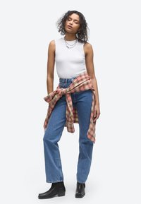 Femme aux cheveux bouclés portant un haut blanc sans manches, un jean bleu, des bottes noires et une chemise à carreaux rouge nouée autour de la taille.