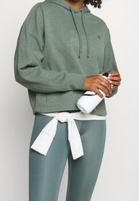 Felpa verde con cappuccio e lacci, dallo stile rilassato. Abbinata a leggings leggeri verdi a vita alta e a un top bianco annodato sopra. Tieni in mano una bottiglia d'acqua.