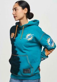 Hoodie teal avec un design en dégradé, des cordons orange et des logos des Miami Dolphins. Comprend une poche kangourou et des accents en tissu texturé.