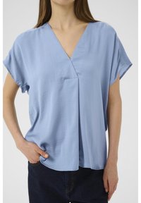 Blusa azzurra chiara con scollatura a V, maniche corte e vestibilità ampia. Presenta una piega frontale e una texture di tessuto liscia.