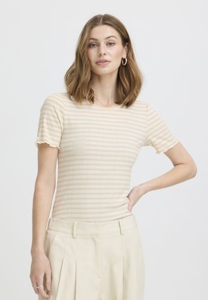 Donna con capelli castani a lunghezza delle spalle che indossa una maglietta a maniche corte beige a righe e pantaloni plissettati color crema, posa con una mano sul fianco.