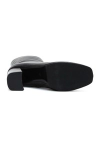 Botte noire montée jusqu'au genou avec un dessus en cuir lisse, un talon bloc rectangulaire et une semelle texturée. Elle présente un bout arrondi et des lignes de design épurées.
