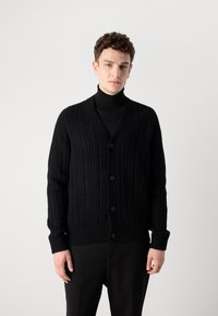 Μαύρο κάρντιγκαν με υφή, ριγωτό σχέδιο, διαθέτει V-λαιμόκοψη, κούμπωμα με κουμπιά και μακριά μανίκια. Φοριέται πάνω από ένα μαύρο φούτερ με στάμπα.