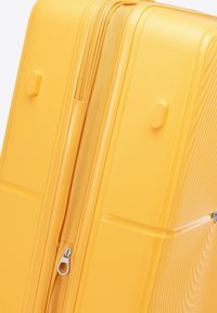 Valigia rigida gialla con superficie testurizzata, chiusura con zip e dettagli modellati. Presenta maniglie laterali e un design elegante.