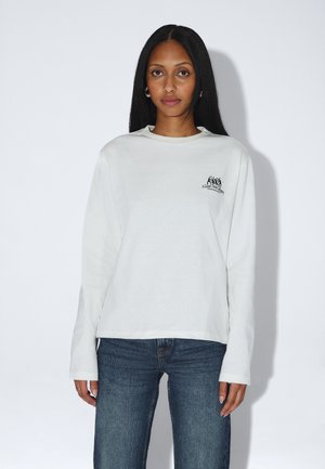 Frau mit langen schwarzen Haaren, die Creolen, ein weißes Langarmshirt mit kleinem schwarzem Textlogo und blaue Jeans trägt, steht vor einem weißen Hintergrund.