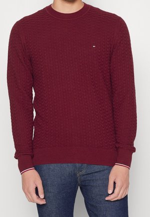 Homme portant un pull bordeaux texturé avec un petit logo sur la poitrine et un jean bleu, debout devant un fond uni clair.