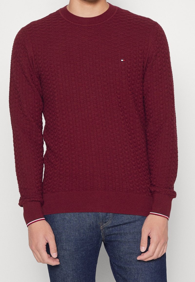 Homme portant un pull bordeaux texturé avec un petit logo sur la poitrine et un jean bleu, debout devant un fond uni clair.