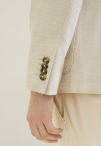 Blazer beige clair avec un motif en chevrons, présentant quatre boutons ronds verts foncés au poignet et une poche visible.