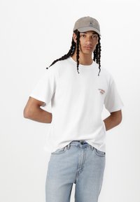 White cotton t-shirt with short sleeves, featuring a small logo in the chest area. Paired with light blue denim jeans and a gray cap.