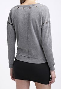 Sudadera gris de manga larga, con detalles de ojales en los hombros y la espalda. Material suave, ajuste holgado y cintura con acabado acanalado.