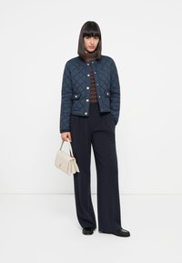 Quiltet navy jakke med gullfarge detaljer, to frontlommer, sammen med en brune stripete turtleneck og vide navy bukser, holder en beige veske.