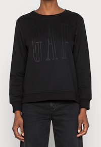 Svart sweatshirt med långa ärmar, rundad nacke, med en stor prägling av "GAP"-logotypen på framsidan. Tillverkad av mjukt tyg med en avslappnad passform.