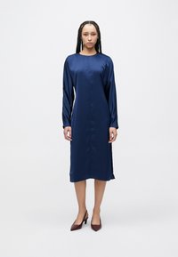 Tengerészkék szatén ruha hosszú ujjakkal, kerek nyakkivágással és oldalsó sliccel. Térdig érő, sima tapintású. Bordó pántos magassarkú cipővel viselve.