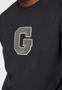 Sweat-shirt noir à col rond avec une grande lettre "G" grise texturée brodée sur la poitrine.