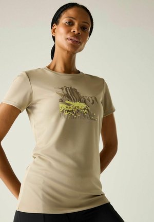 Kobieta ubrana w beżowy dopasowany T-shirt z grafiką natury przedstawiającą skały i żółte kwiaty, stojąca na tle jednolitego jasnego tła.