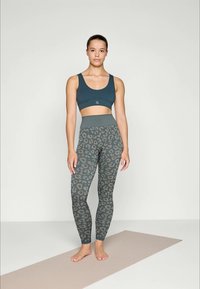 Ung kvinne står barfot på en beige yogamatte iført mørk teal sports-BH og høyt liv leggings med grå leopardmønster.