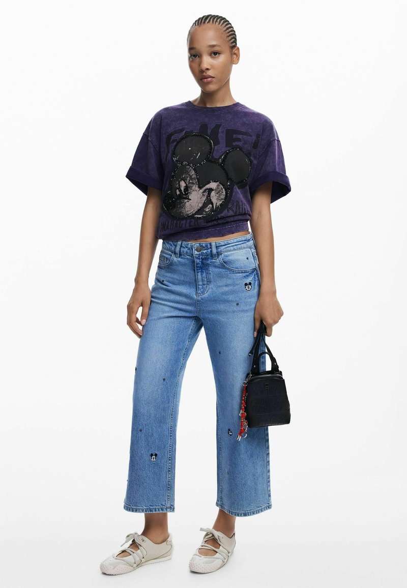 T-shirt corta viola con grafica di Topolino, abbinata a jeans larghi azzurri con piccole toppe. Accessori includono una piccola borsa nera.