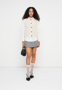 Beige strikket vest med knapper over en hvit krage skjorte, grå plissert skjørt, gjennomsiktige hvite sokker, og svarte pumps med sløyfe. Svart håndveske.