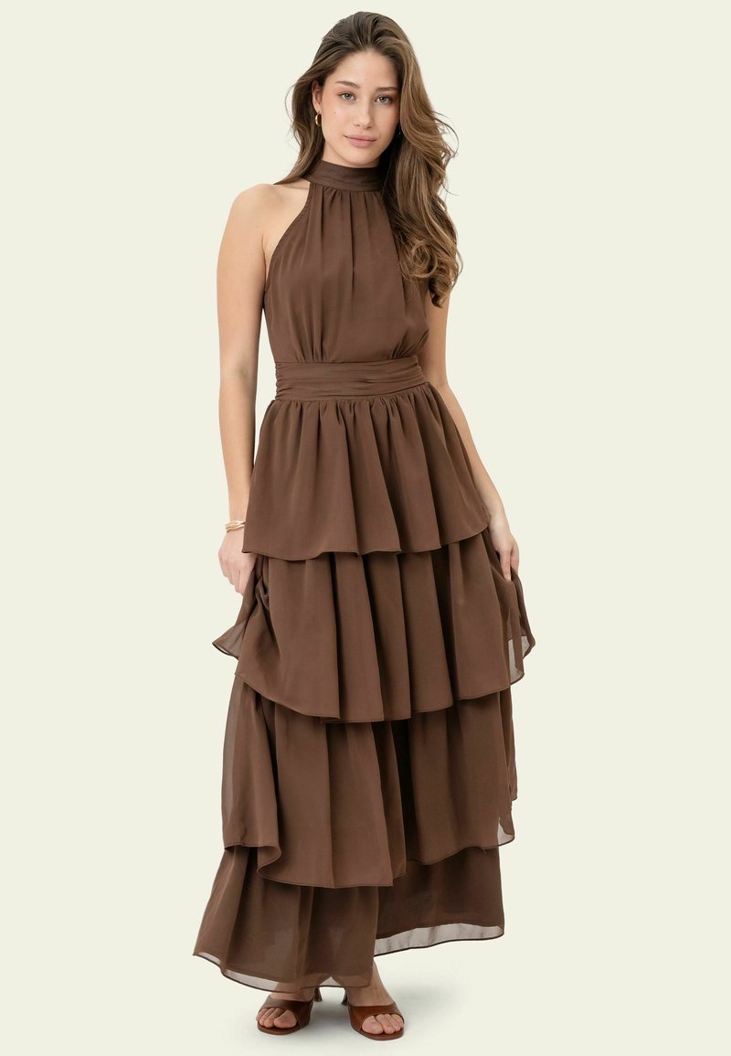 Jeune femme debout portant une robe maxi marron à volants de type halter-neck et des sandales marron sur un fond clair uni.