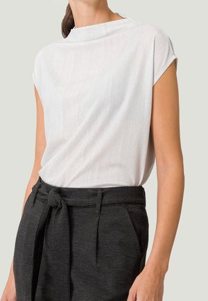 Kobieta w białej teksturowanej bluzce z krótkim rękawem, wpuszczonej w czarne spodnie paperbag z wysokim stanem i wiązanym paskiem.