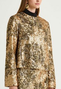 Haut à sequins dorés avec une surface texturée, présentant un col montant et des manches longues. Motifs floraux décoratifs et ornements présents.