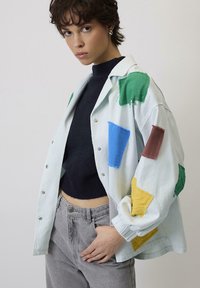 Svijetloplava oversized košulja s raznim tekstilnim zakrpama u zelenoj, plavoj, smeđoj i žutoj boji. Ima metalne gumbe i opušteni kroj.