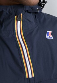 Marineblå letvægtsjakke med halvmånedalsdesign, farverige lodrette striber og K-Way-logo på brystet. Justerbar kædetræk ved halsen.