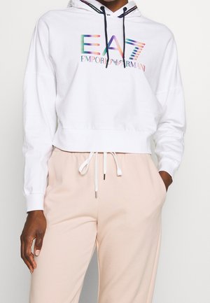 Persoon die een witte cropped hoodie draagt met het EA7 Emporio Armani-logo in regenboogkleuren, gecombineerd met beige broek met trekkoord.