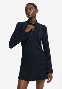 Rochie de culoare albastru marin, cu mâneci lungi, guler croiel, și dungi verticale albe. Prezintă o croială ajustată și ușoară strângere pe laterale.